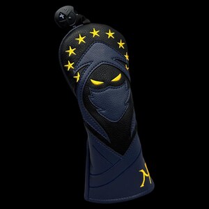 May include: A navy and black golf club head cover with a ninja design. The cover features a ninja face with yellow eyes and stars, and the letter "N" in yellow. The cover has a black top with a clip.