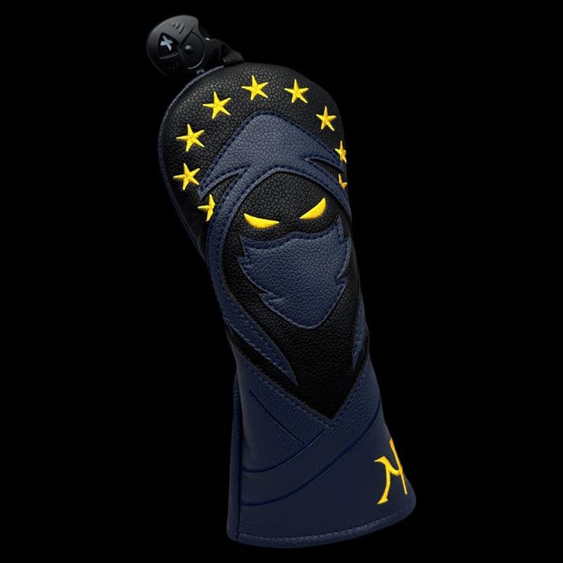 May include: A navy and black golf club head cover with a ninja design. The cover features a ninja face with yellow eyes and stars, and the letter "N" in yellow. The cover has a black top with a clip.