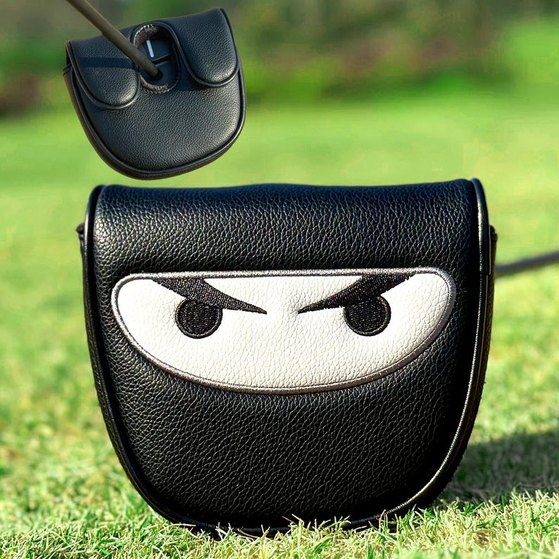 Może przedstawiać: Czarny sk&oacute;rzany pokrowiec na putter golfowy z motywem twarzy ninja. Pokrowiec ma białą twarz z czarnymi haftowanymi oczami i srebrnym obramowaniem. Pokrowiec na putter jest pokazany na tle zielonej trawy.