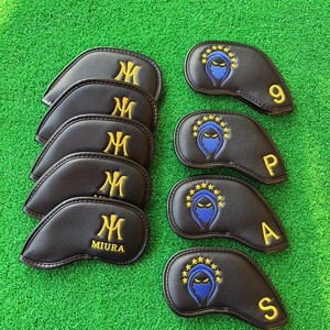 May include: A collection of black golf club head covers. Some feature a gold "M" logo, while others display a blue ninja design with gold stars and the numbers 9, P, A, and S in gold. The covers are arranged on a green surface.