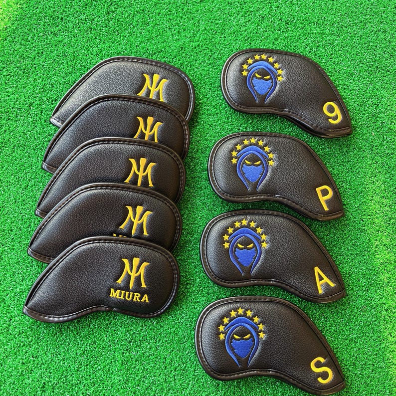May include: A collection of black golf club head covers. Some feature a gold "M" logo, while others display a blue ninja design with gold stars and the numbers 9, P, A, and S in gold. The covers are arranged on a green surface.