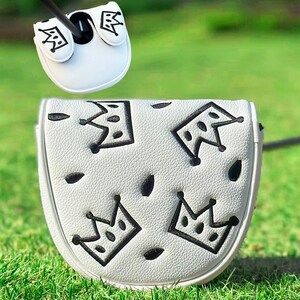 Pu&ograve; includere: Copertura per putter da golf bianca con disegni di corone nere. La copertura &egrave; realizzata in un materiale strutturato e presenta una chiusura magnetica. Il design include diverse illustrazioni di corone nere e piccole forme ovali nere. La copertura del putter &egrave; mostrata su una superficie erbosa verde.