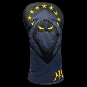 May include: A navy blue and black golf club head cover with a yellow embroidered design. The design features a hooded figure with yellow eyes and stars. The initials "NA" are embroidered in yellow.