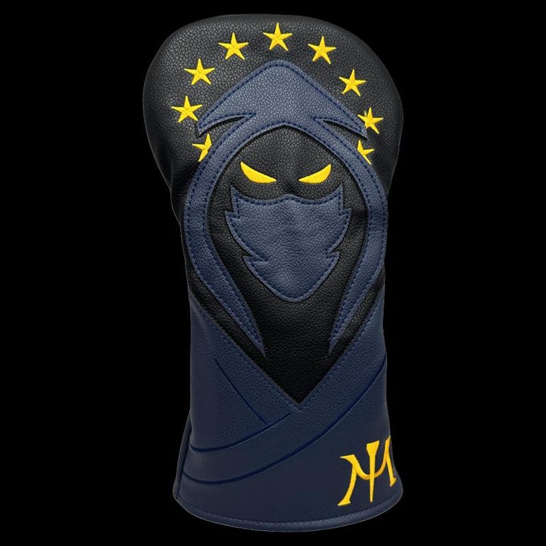 May include: A navy blue and black golf club head cover with a yellow embroidered design. The design features a hooded figure with yellow eyes and stars. The initials "NA" are embroidered in yellow.