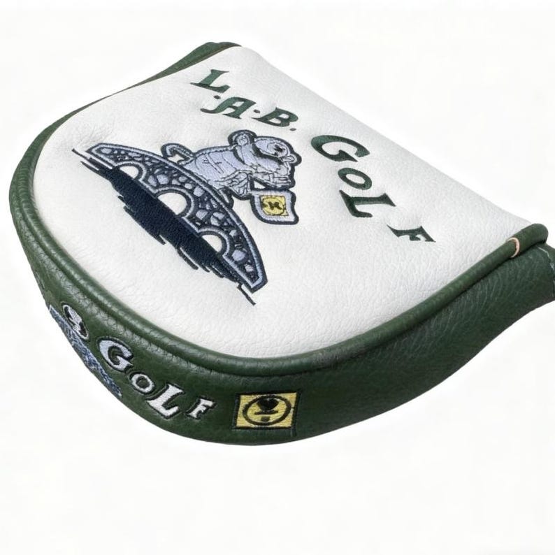 May include: White and green golf club head cover with embroidered design. The cover features the text "LAB GOLF" and a graphic of a bridge and a bear. The cover has a green border and a white top.