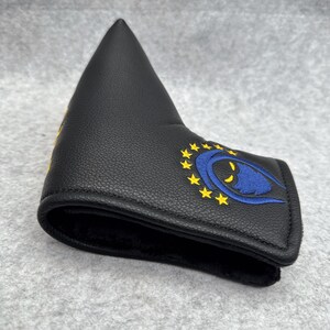 May include: Black golf putter head cover with a blue and yellow embroidered design. The design features a blue eye shape surrounded by yellow stars. The cover is made of a textured material.