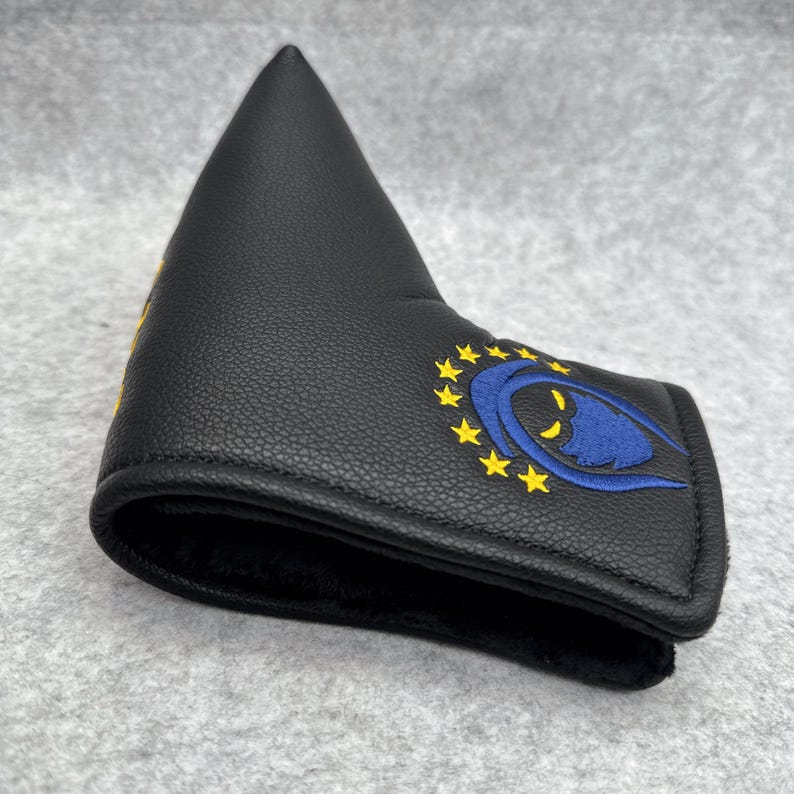 May include: Black golf putter head cover with a blue and yellow embroidered design. The design features a blue eye shape surrounded by yellow stars. The cover is made of a textured material.