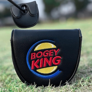 Może przedstawiać: Czarny pokrowiec na gł&oacute;wkę puttera golfowego z napisem "Bogey King" wyhaftowanym na czerwono i ż&oacute;łto, z niebieską obw&oacute;dką. Pokrowiec wykonany jest z teksturowanego materiału i jest pokazany na zielonej trawie.
