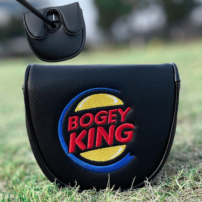 Może przedstawiać: Czarny pokrowiec na gł&oacute;wkę puttera golfowego z napisem "Bogey King" wyhaftowanym na czerwono i ż&oacute;łto, z niebieską obw&oacute;dką. Pokrowiec wykonany jest z teksturowanego materiału i jest pokazany na zielonej trawie.