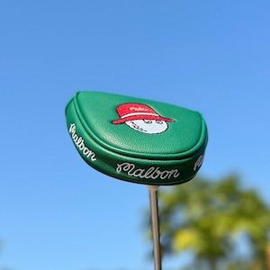 May include: A green golf club head cover with the word "Malbon" embroidered in white. The cover features an embroidered cartoon golf ball wearing a red hat. The background is a clear blue sky.