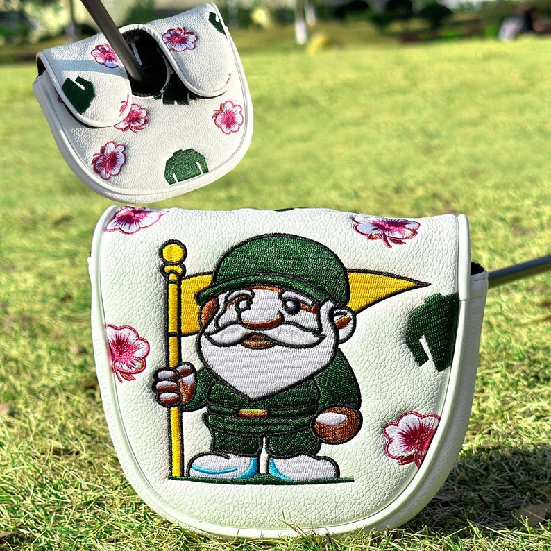 Puede incluir: Dos fundas blancas para la cabeza del palo de golf. Una funda presenta un gnomo de dibujos animados sosteniendo una bandera, la otra tiene un estampado floral. Ambas fundas tienen un fondo blanco con detalles en verde y rosa.