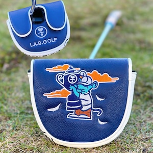 Pu&ograve; includere: Due copricapo per mazze da golf blu navy con finiture bianche. Un copricapo ha il testo "L.A.B. GOLF" e un simbolo a forma di cuore. L'altro copricapo presenta un personaggio dei cartoni animati che tiene un trofeo, con nuvole arancioni e dettagli bianchi.