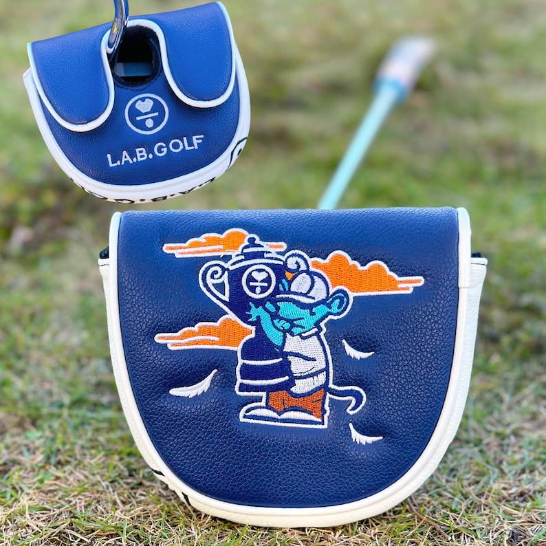 Pu&ograve; includere: Due copricapo per mazze da golf blu navy con finiture bianche. Un copricapo ha il testo "L.A.B. GOLF" e un simbolo a forma di cuore. L'altro copricapo presenta un personaggio dei cartoni animati che tiene un trofeo, con nuvole arancioni e dettagli bianchi.