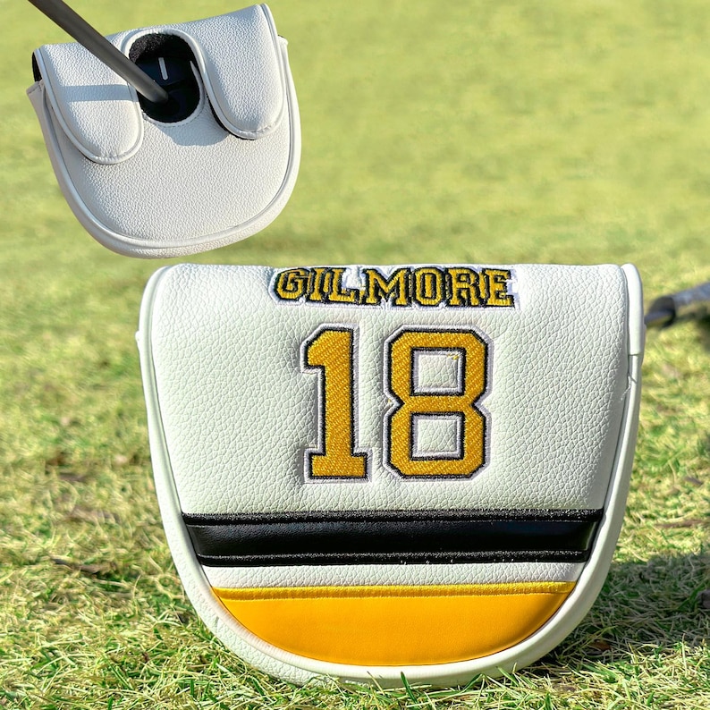 Pu&ograve; includere: Copertura bianca per la testa della mazza da golf con il nome "GILMORE" in lettere dorate e il numero "18" in oro con contorno nero. La copertura presenta un motivo a strisce nere e gialle nella parte inferiore. Una mazza da golf &egrave; inserita nella copertura.
