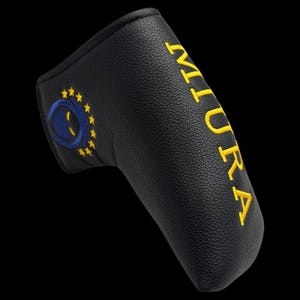 May include: Black golf putter cover with yellow embroidered text "MIURA" and a blue and yellow emblem. The cover is made of textured material and is designed to protect the golf club head.