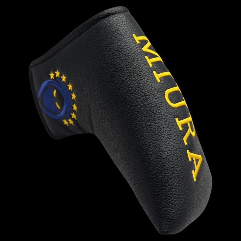 May include: Black golf putter cover with yellow embroidered text "MIURA" and a blue and yellow emblem. The cover is made of textured material and is designed to protect the golf club head.