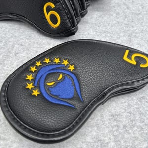 May include: Black golf club head covers with yellow embroidered numbers 5 and 6. One cover features a blue and yellow emblem with a star design. The covers are made of a textured material.