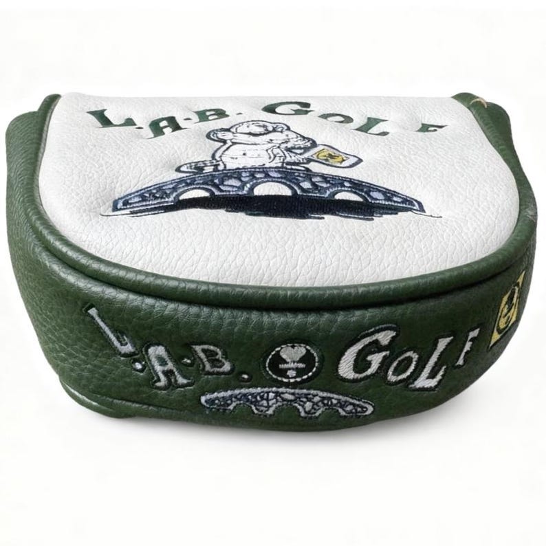 May include: A golf putter head cover with a white top and green sides. The top features the text "LAB GOLF" and a cartoon image of a bear on a bridge. The sides also have the text "LAB GOLF".