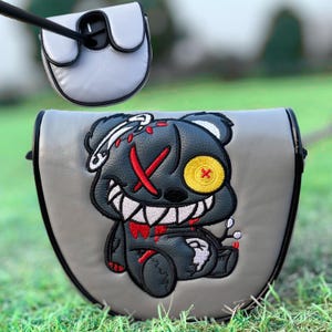 May include: A grey golf club head cover featuring an embroidered cartoon bear design in black and red. The bear has a yellow eye, a red 'X' over its eye, and a safety pin. The cover is made of a grey material with black trim.