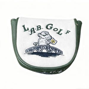 May include: A white and green golf club head cover with the text "LAB GOLF" and a cartoon monkey holding a flag. The cover has a green border and a white center with a bridge design.