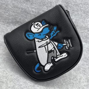 Puede incluir: Funda para la cabeza del palo de golf de cuero negro con un ratón de dibujos animados bordado con un atuendo y sombrero blancos, sosteniendo una pesa. El ratón es azul con detalles blancos. La funda tiene forma curva.