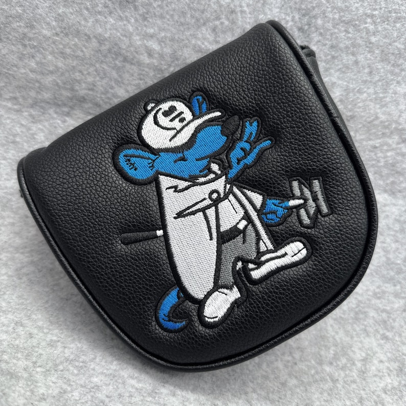 May include: Black leather golf club head cover with an embroidered cartoon mouse in a white outfit and hat, holding a dumbbell. The mouse is blue with white accents. The cover has a curved shape.
