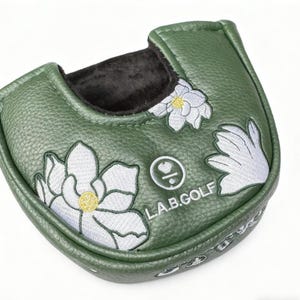 May include: A green golf putter cover with white embroidered floral designs and the text "L.A.B.GOLF". The cover has a textured surface and a black interior lining. The floral design includes yellow accents.