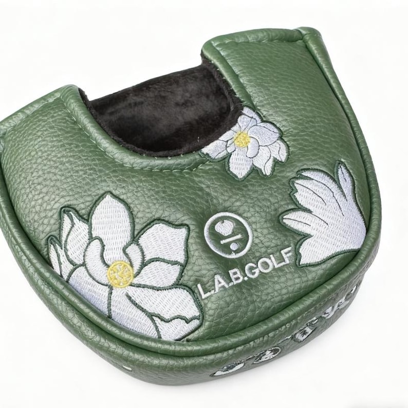 May include: A green golf putter cover with white embroidered floral designs and the text "L.A.B.GOLF". The cover has a textured surface and a black interior lining. The floral design includes yellow accents.