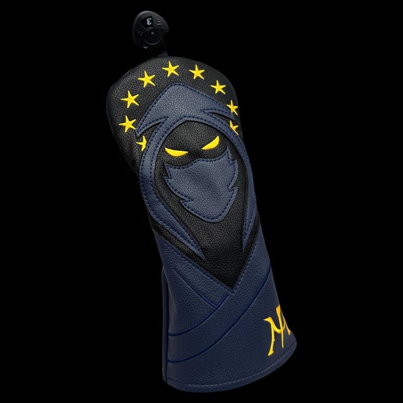 May include: A navy blue and black golf club head cover with a stylized ninja design. The cover features yellow star accents and yellow eyes. The letter "M" is embroidered in yellow on the side.