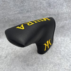 May include: Black golf club head cover with yellow embroidered text reading "MITRA" on top and "M" on the side. The cover is made of a textured material.