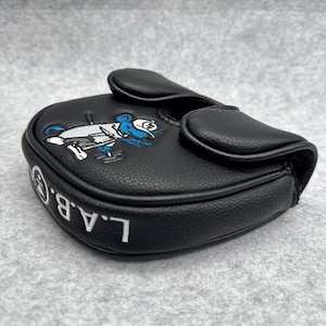 May include: Black golf putter cover with a cartoon character design in white and blue. The cover has the letters "L.A.B.G." in white. The cover is made of a textured material.