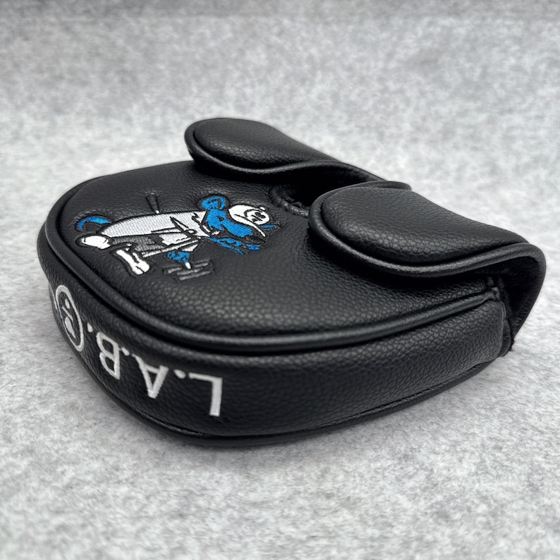 May include: Black golf putter cover with a cartoon character design in white and blue. The cover has the letters "L.A.B.G." in white. The cover is made of a textured material.