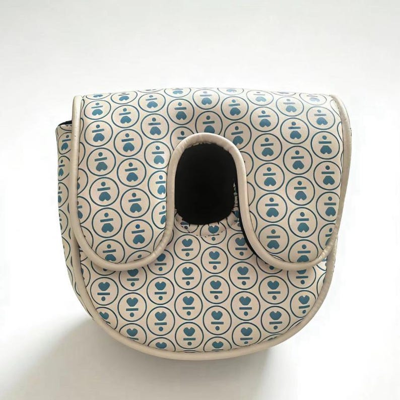 May include: A cream-colored bag with a repeating pattern of blue hearts and lines inside circles. The bag has a rounded shape with a black opening and a beige trim.