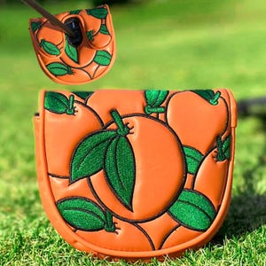 Puede incluir: Funda para putter de palo de golf naranja con dise&ntilde;o de hojas verdes. La funda est&aacute; hecha de un material similar al cuero y presenta un color naranja vibrante con hojas verdes bordadas. El dise&ntilde;o se repite en la cubierta superior.
