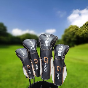 Puede incluir: Un juego de fundas para palos de golf en blanco y negro con la marca "PING" en letras blancas. Las fundas tienen detalles en naranja y el nombre del modelo "G400". El fondo muestra un campo de golf verde bajo un cielo azul.
