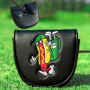 Może przedstawiać: Czarny sk&oacute;rzany pokrowiec na putter golfowy z kresk&oacute;wkowym hot dogiem. Hot dog nosi zieloną czapkę i okulary przeciwsłoneczne, trzymając torbę na kije golfowe. Na tle zielonej trawy.