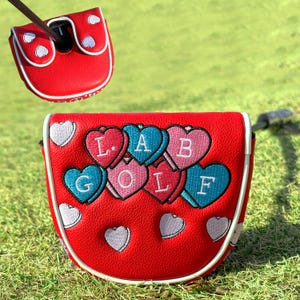Puede incluir: Funda roja para putter de golf con ribete blanco y detalles de coraz&oacute;n. La funda presenta el texto "LAB GOLF" en dise&ntilde;os coloridos en forma de coraz&oacute;n. Se muestra una funda a juego arriba, con un palo de golf insertado.