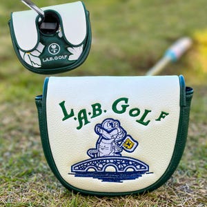 Pu&ograve; includere: Copertura per la testa del putter da golf bianca e verde. La copertura presenta il testo "L.A.B. Golf" e un alligatore dei cartoni animati ricamato che tiene una bandiera. La copertura &egrave; su una superficie erbosa verde.