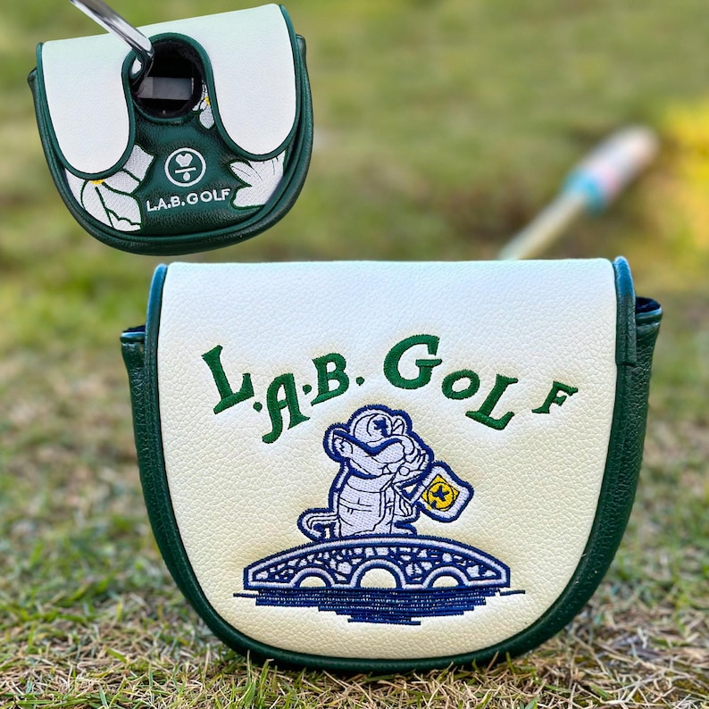 Pu&ograve; includere: Copertura per la testa del putter da golf bianca e verde. La copertura presenta il testo "L.A.B. Golf" e un alligatore dei cartoni animati ricamato che tiene una bandiera. La copertura &egrave; su una superficie erbosa verde.