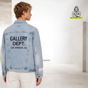 Op de afbeelding: Lichtblauwe spijkerjas met de tekst "GALLERY DEPT. LOS ANGELES, CA." op de rug. De jas heeft een klassiek ontwerp met knopen en zakken. Het model draagt een witte broek.