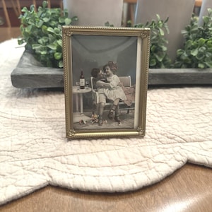 May include: A rectangular gold-toned metal picture frame with a decorative beaded edge and floral corner accents. The frame holds a vintage-style print depicting two children sitting on a bench with a small table, a bottle, and glasses.