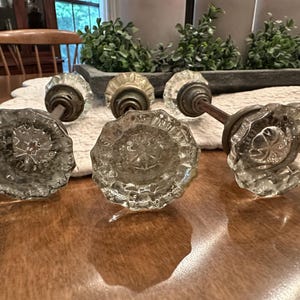 May include: Three clear glass door knobs with a faceted, multi-sided design and a star-shaped pattern in the center. Each knob is attached to a dark, weathered metal spindle and base, showing a classic, functional hardware construction.