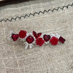 May include: A silver barrette featuring red heart and rose-shaped gems, along with square and teardrop-shaped red gems. The barrette is adorned with small, clear crystals and pearls.