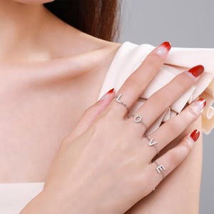 May include: A set of four silver-toned rings worn on four fingers, each featuring a single letter charm spelling out the word LOVE. Each letter is encrusted with small, clear, sparkling stones, and the rings are connected by thin silver chains.