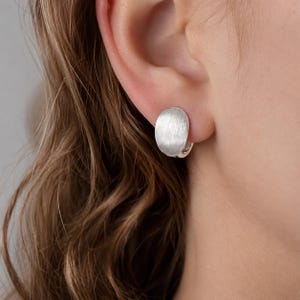 Pode incluir: Um brinco de tom prateado com um design de disco circular de metal escovado. O brinco tem um acabamento fosco e liso com uma textura de linhas finas visível em toda a superfície, preso com um pequeno fecho de argola no lóbulo.