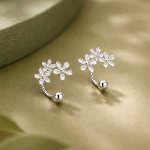 Könnte beinhalten: Ein Paar silberfarbene Ohrringe mit einem geschwungenen Stabdesign, das mit drei kleinen blumenförmigen Anhängern verziert ist. Jede Blume hat eine texturierte, funkelnde Oberfläche auf den Blütenblättern und eine glatte, polierte Mitte.