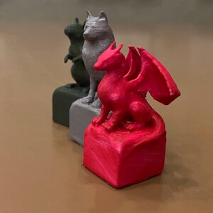 Puede incluir: Tres peque&ntilde;as figuras de animales: una ardilla verde, un lobo gris y un drag&oacute;n rojo con alas extendidas. Cada figura est&aacute; montada sobre una base cuadrada. El drag&oacute;n rojo es el punto focal.