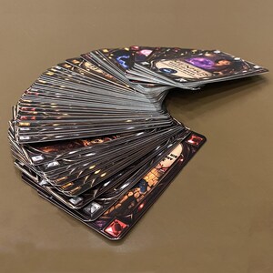 Puede incluir: Un mazo de cartas abanicado, con bordes oscuros y ornamentados y detalles de joyas. Las cartas tienen ilustraciones y texto detallados, con un tema de fantas&iacute;a. La carta superior muestra la palabra "MENHUS".