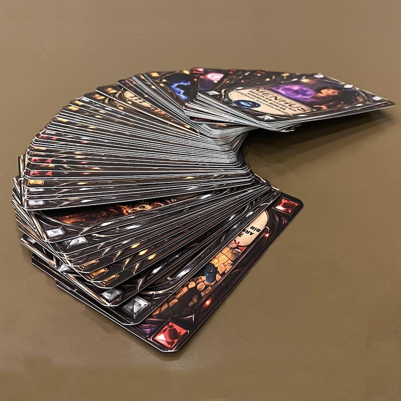 Puede incluir: Un mazo de cartas abanicado, con bordes oscuros y ornamentados y detalles de joyas. Las cartas tienen ilustraciones y texto detallados, con un tema de fantas&iacute;a. La carta superior muestra la palabra "MENHUS".