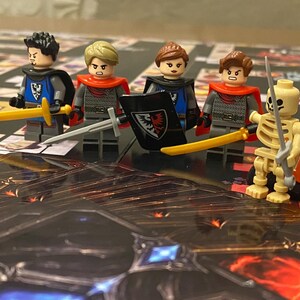 Puede incluir: Una configuraci&oacute;n de juego de mesa con minifiguras de Lego de caballeros y un guerrero esqueleto, dados y piezas de juego. Los caballeros est&aacute;n armados con espadas y escudos, y los dados son naranjas. El tablero del juego tiene un tema de fantas&iacute;a.